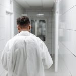Profissional de saúde de jaleco caminha por um corredor iluminado de hospital, visto de costas.