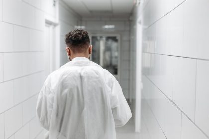Profissional de saúde de jaleco caminha por um corredor iluminado de hospital, visto de costas.