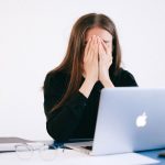 Mulher demonstra estresse e sobrecarga sentada à mesa diante de um laptop, em ambiente de trabalho.