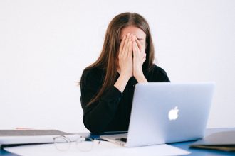 Mulher demonstra estresse e sobrecarga sentada à mesa diante de um laptop, em ambiente de trabalho.