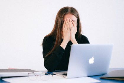 Mulher demonstra estresse e sobrecarga sentada à mesa diante de um laptop, em ambiente de trabalho.